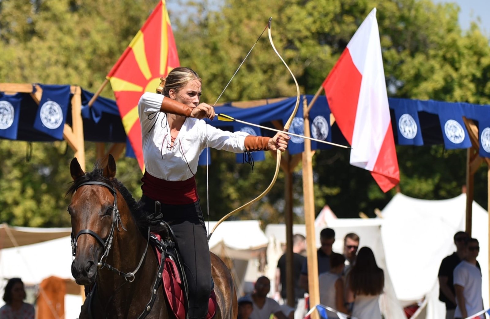 A rider drawing a traditional bow in the saddle, with flags flying over the course behind her