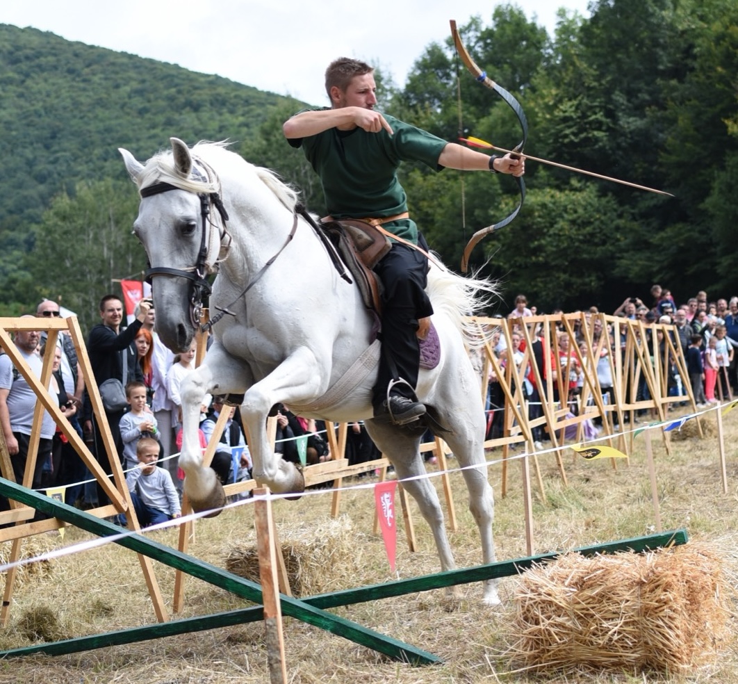 A horseback archer on a grey horse drawing his bow as the horse leaps along the course