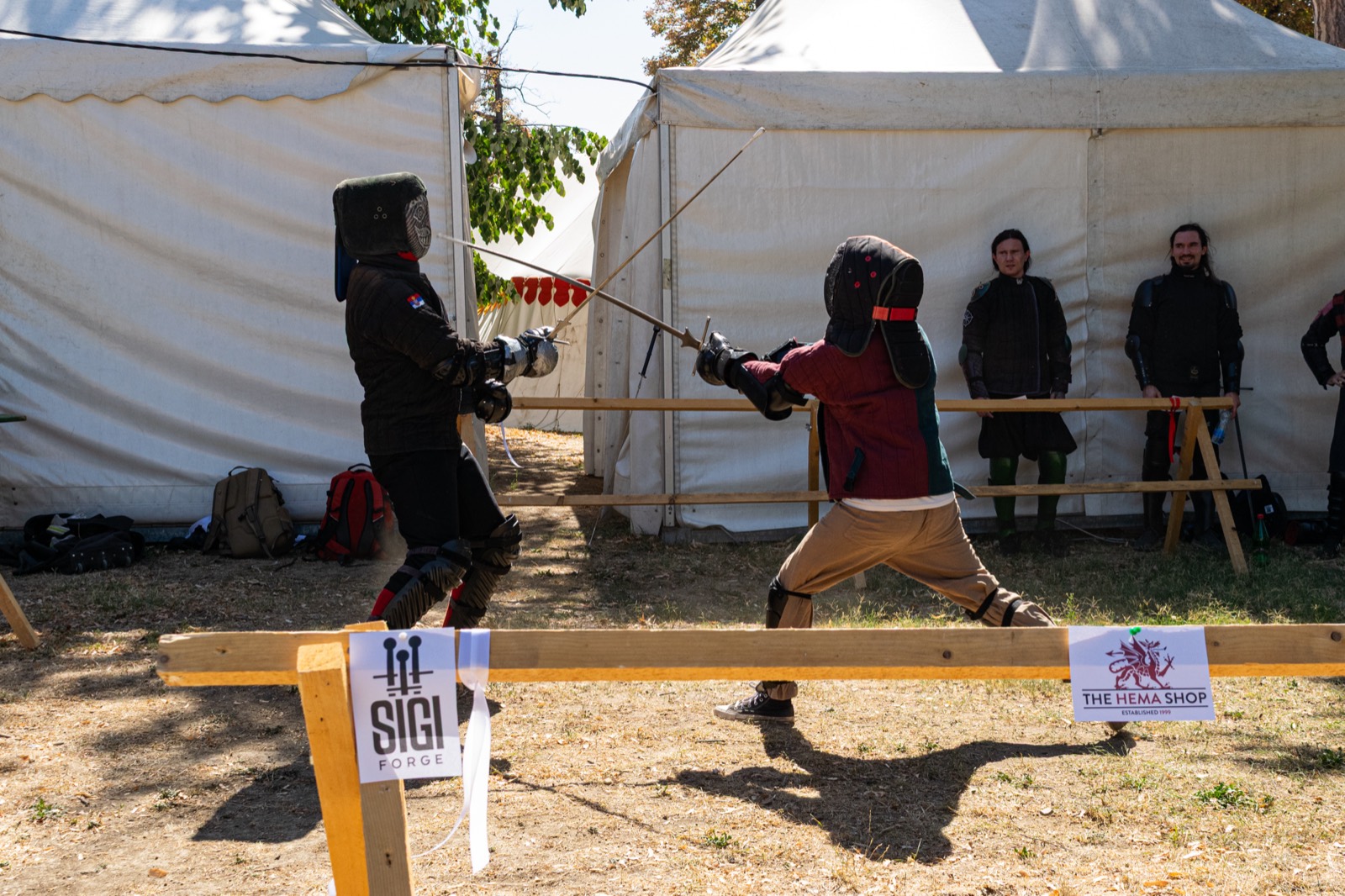 Two fencers crossing longswords in a ring lined with tents, with SIGI Forge and The HEMA Shop banners on the rail